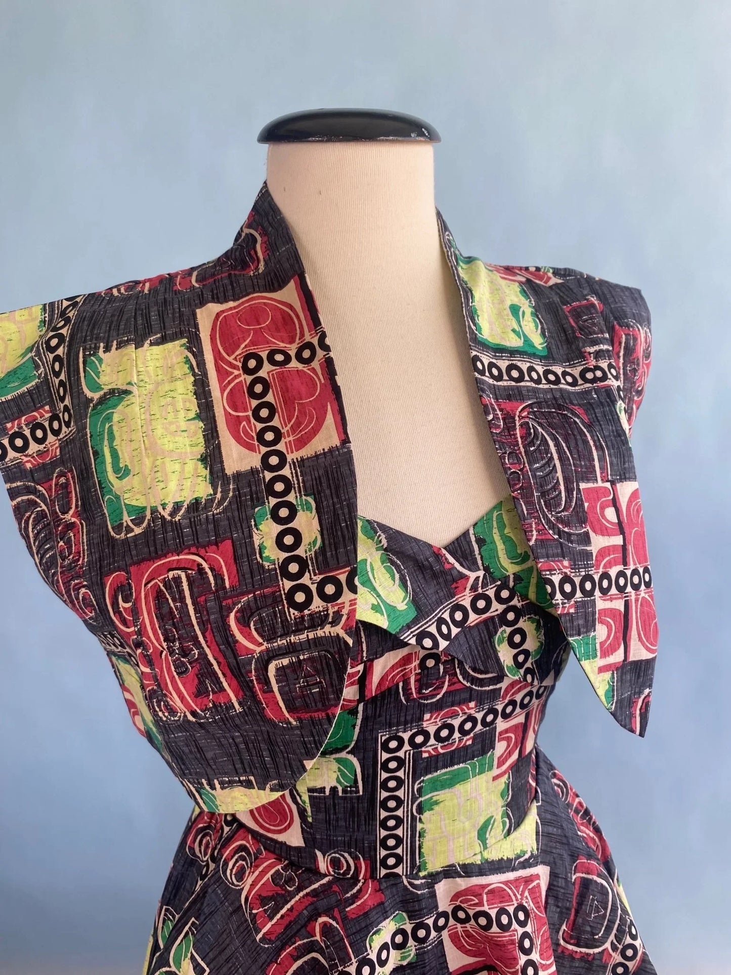 Vintage-inspired dress with bold geometric print in red, green, black, and cream, featuring structured shoulders and wrap neckline.